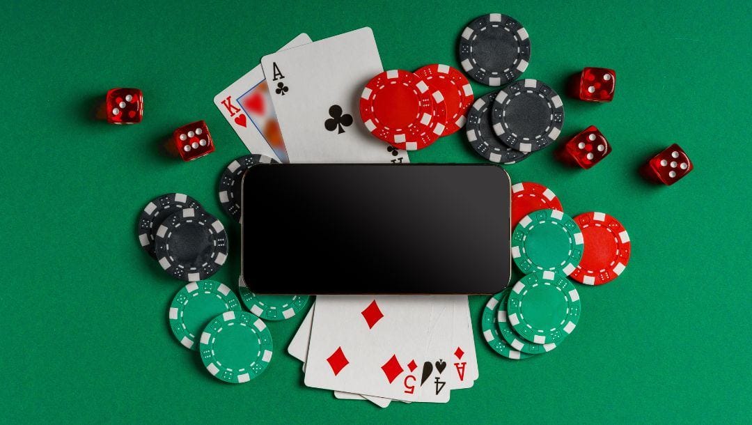 An online poker concept image showing a smartphone on a green felt poker table surrounded by playing cards, dice, and poker chips.