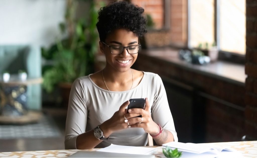 Cheerful woman sitting at table using cellphone with laptop and book on table