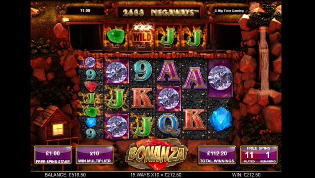 A screenshot of the free spins mode in the Bonanza Megaways slot. The game is set in a mining quarry that’s bathed in red light. Visible on the reels are a variety of slot symbols, including red, green, and blue gems, diamonds, and the A, K, Q, and J. To the left of the reels is a mining shack, and on the right is a spinning water wheel. Above the reels is an extra row of symbols. Each symbol is in a mining cart.