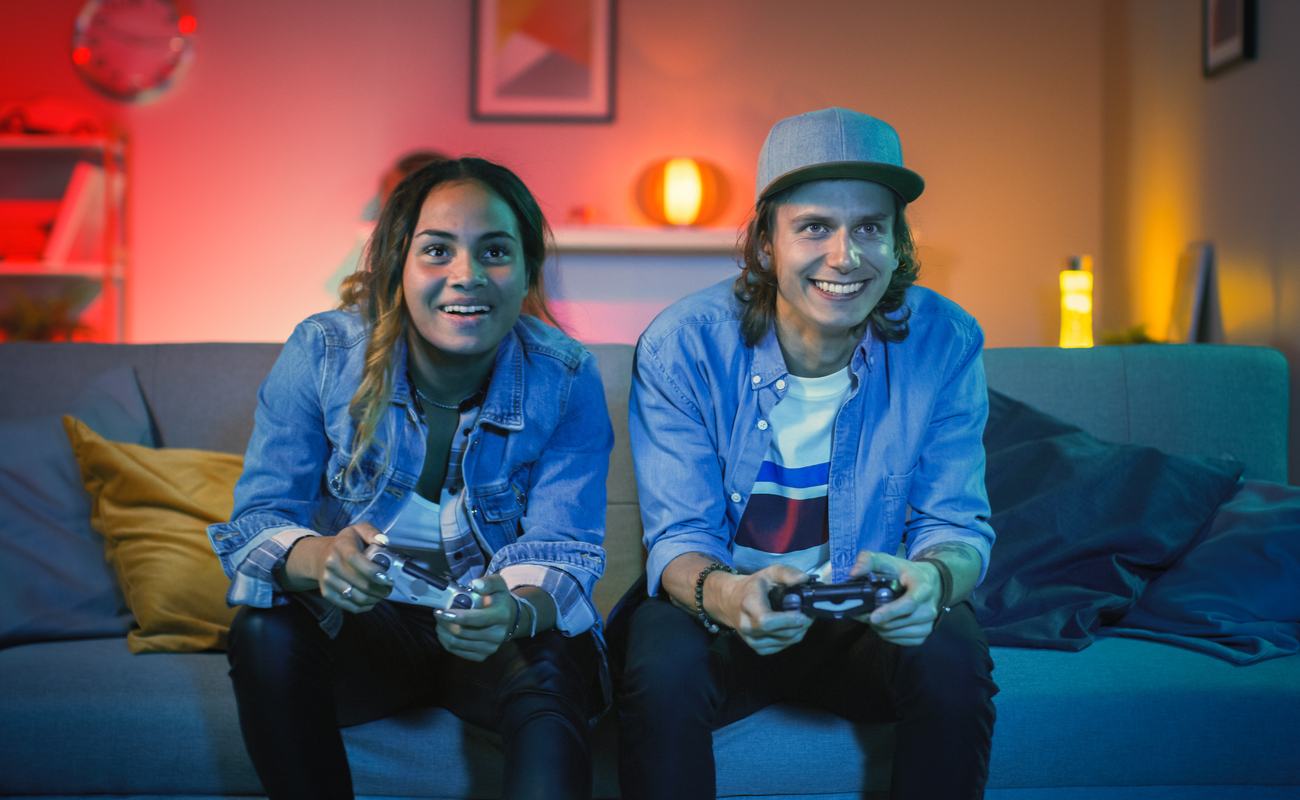 A smiling young couple sitting on a couch playing video games together in a cozy, colorful living room.