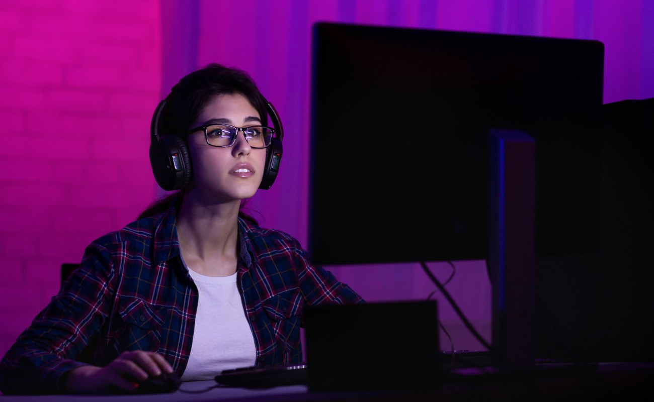 A woman wearing headphones and glasses focused on a computer screen in a purple-lit room.