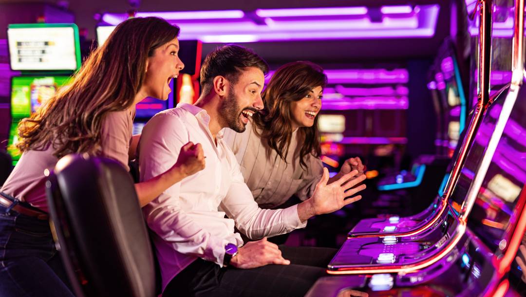 Two women and a man are celebrating in front of slot machines at a casino.