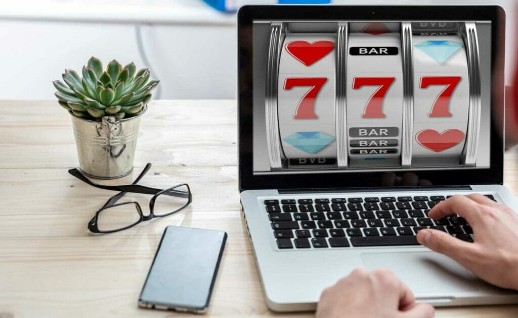 A man plays online slots on a laptop with a mobile phone, spectacles and a succulent plant beside him.