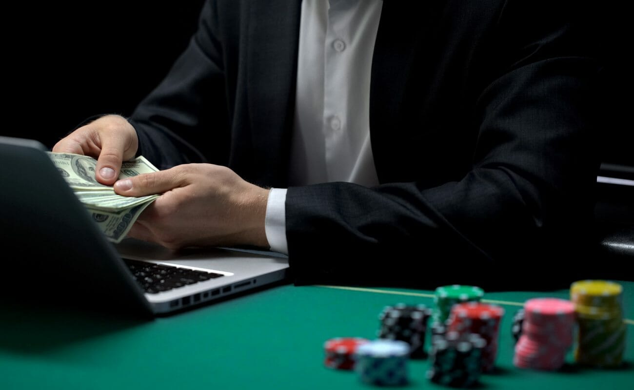 An online poker player with money in his hand in front of a laptop and casino chips.
