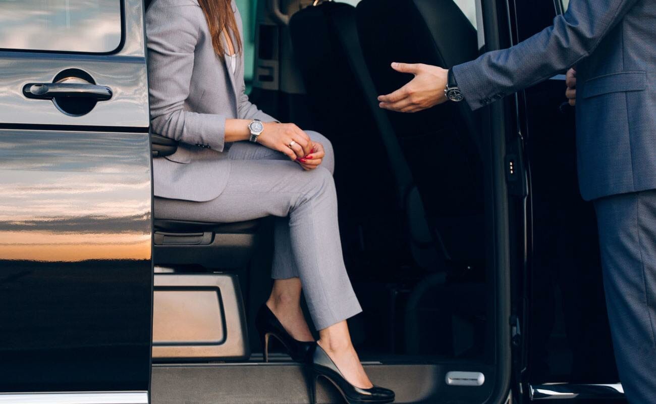 A driver helps a person in a suit and heels out of a car.