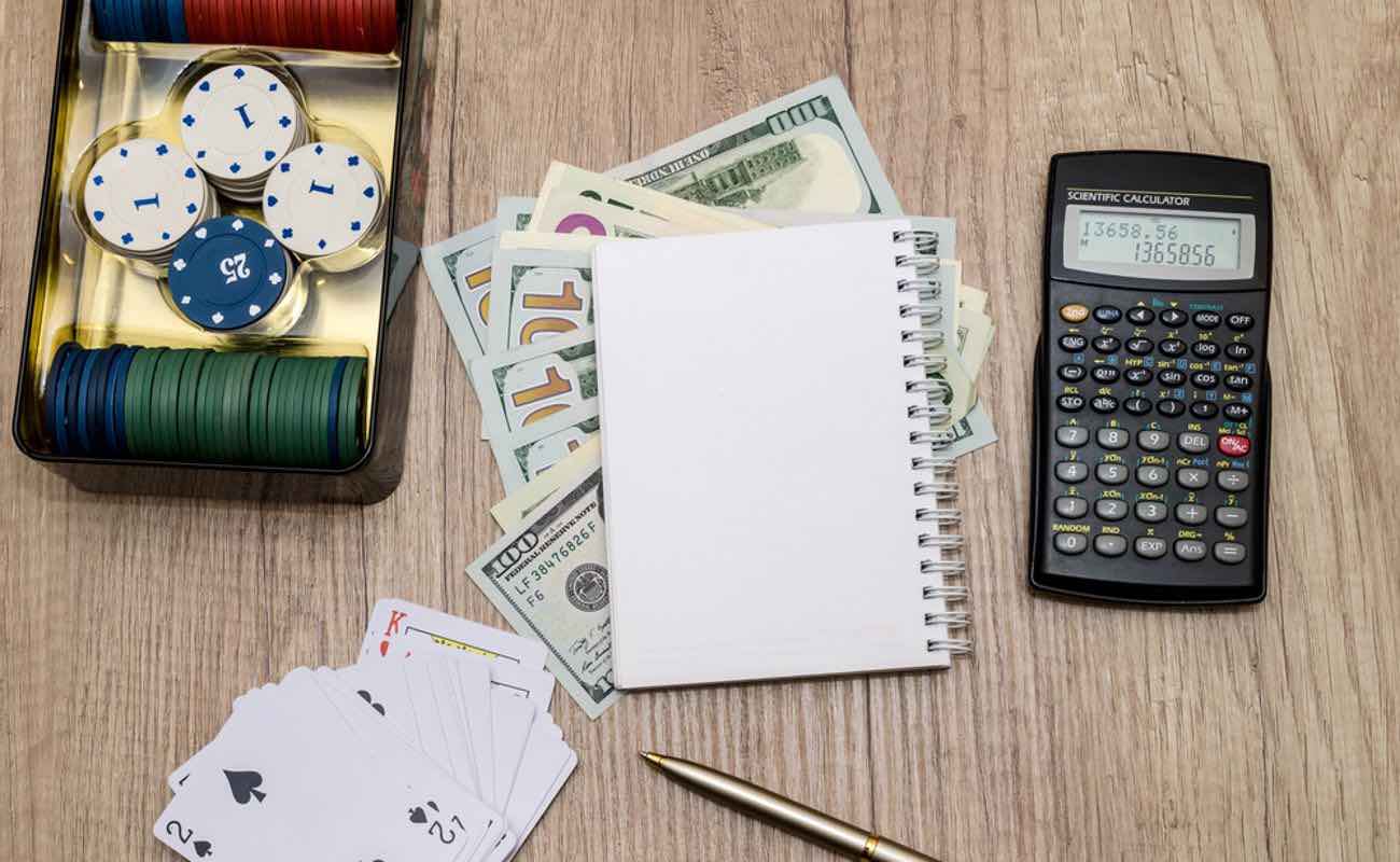A notebook, money, calculator, casino chips and playing cards on a table.