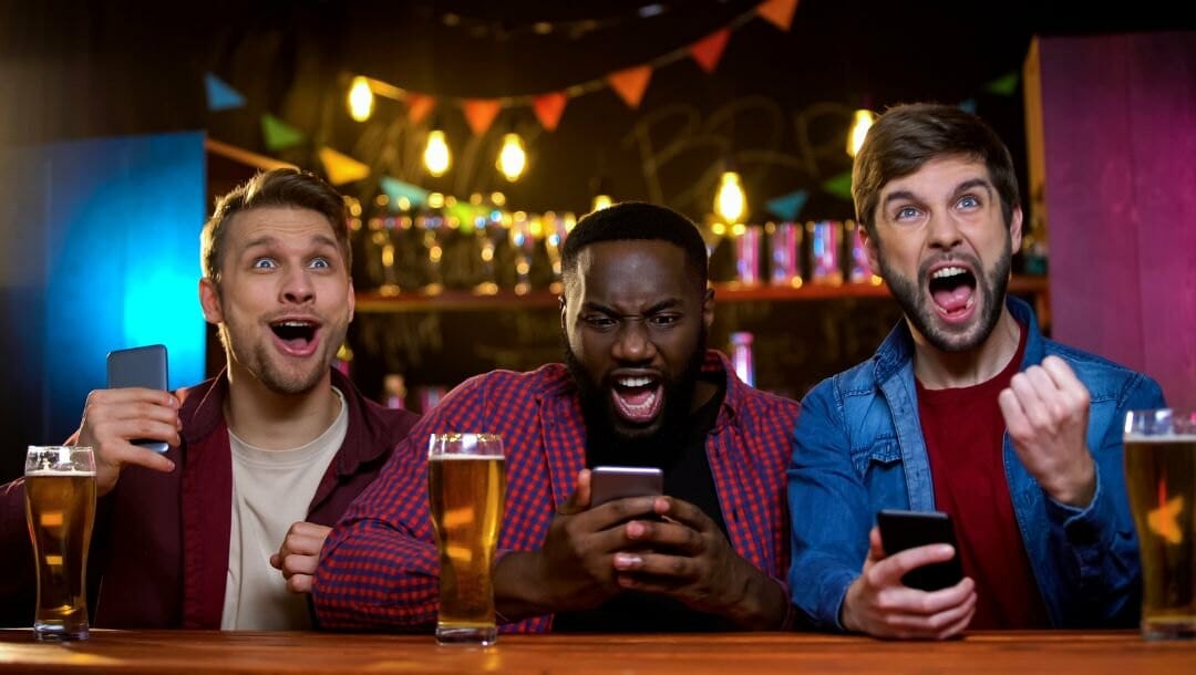 three excited males gripping their phones while cheering for their sports team at a bar with a beer in front of each man