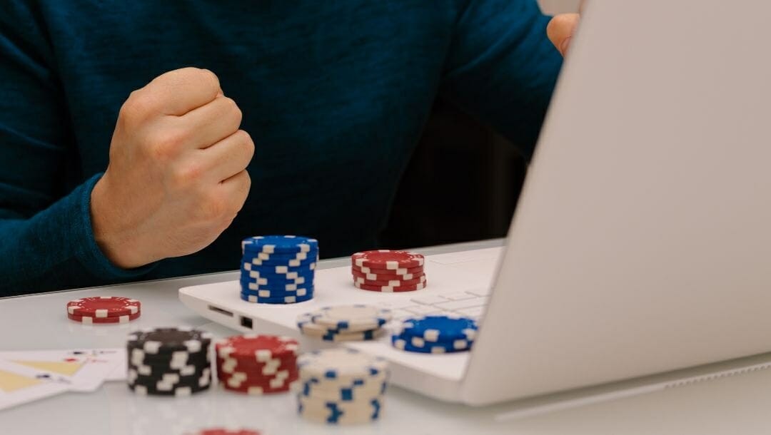 Body image, man cheering while online gambling on a laptop, poker chips stacked around and three ace cards fanned out on the table