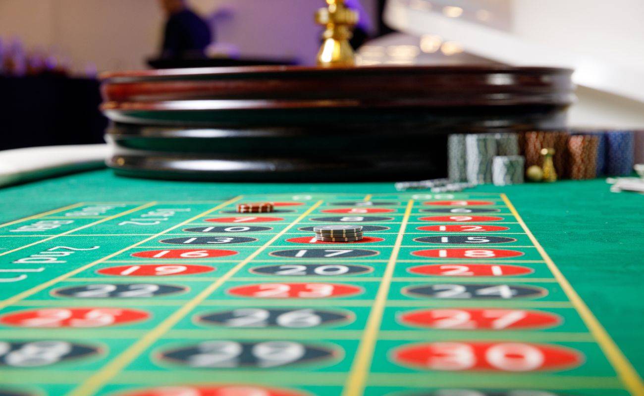 low angle shot of a roulette table with poker chip bets stacked on it with the roulette wheel in the back next to stacks of poker chips