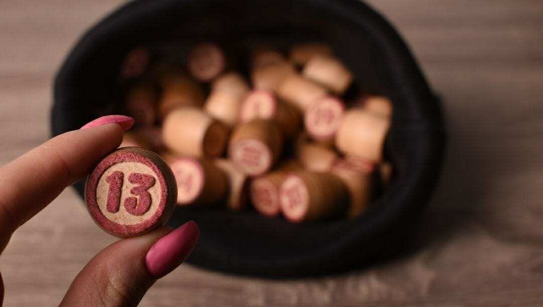 unlucky number 13 wooden bingo number held in a woman’s fingers, her fingernails are painted pink, container full of other bingo numbers in the background.
