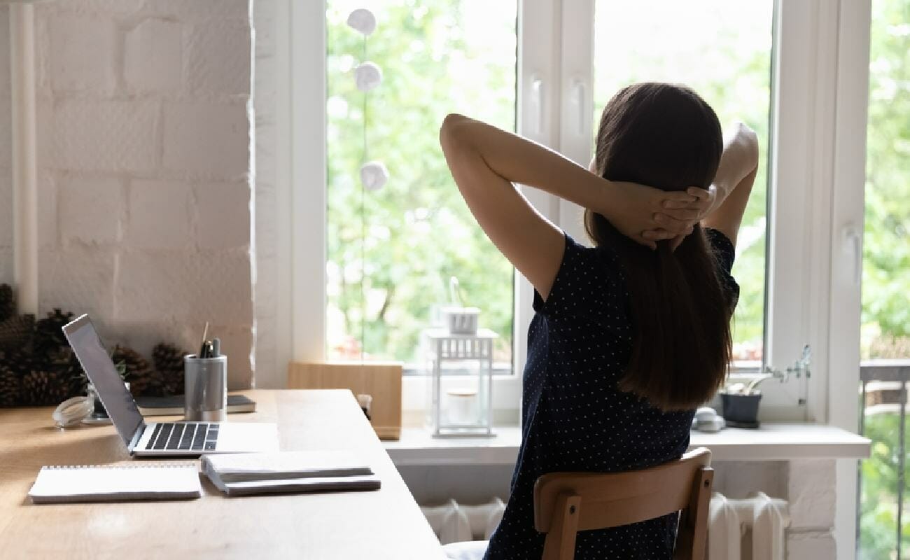 A woman takes a break at her laptop and puts her hands behind her head