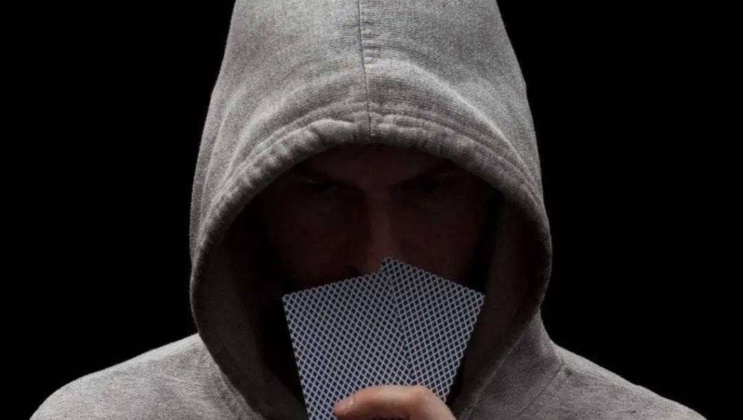 A poker player in a hoodie holding a pair of cards in front of his face.