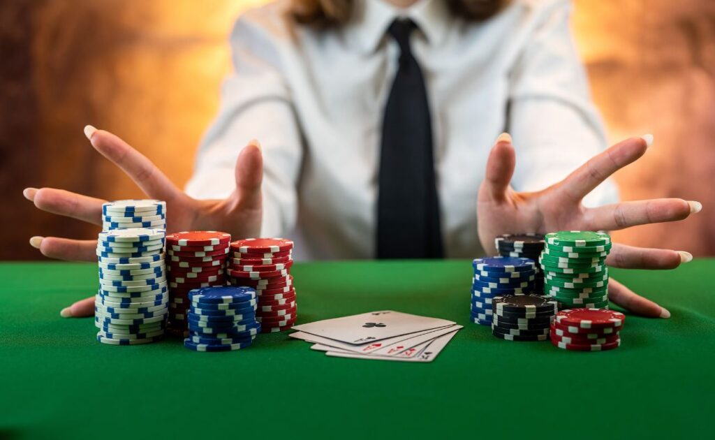 a woman pushes stacks of poker chips forward with both hands on a green poker table with playing cards face up between the stacks