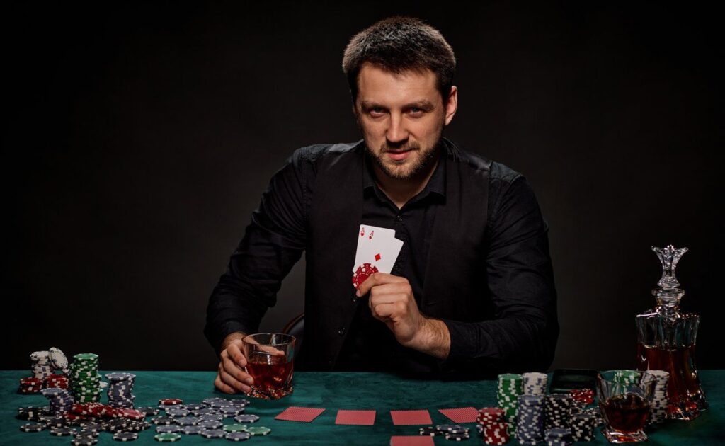 A man holding up two aces and two poker chips in the same hand. He holds his drink in the other while sitting in front of a poker table.