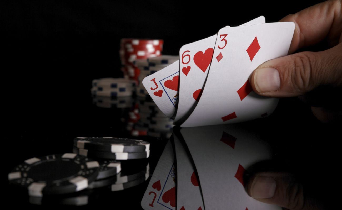 a person checks their three hole cards, a jack and six of hearts and a three of diamonds, on a reflective black surface with poker chips stacked to the side and in the blurred background