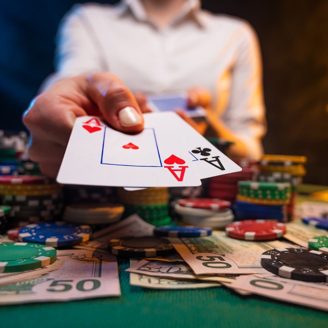 A person seated at a poker table, presenting an Ace of Hearts and an Ace of Clubs in hand, surrounded by a table of money and neatly arranged casino chips.