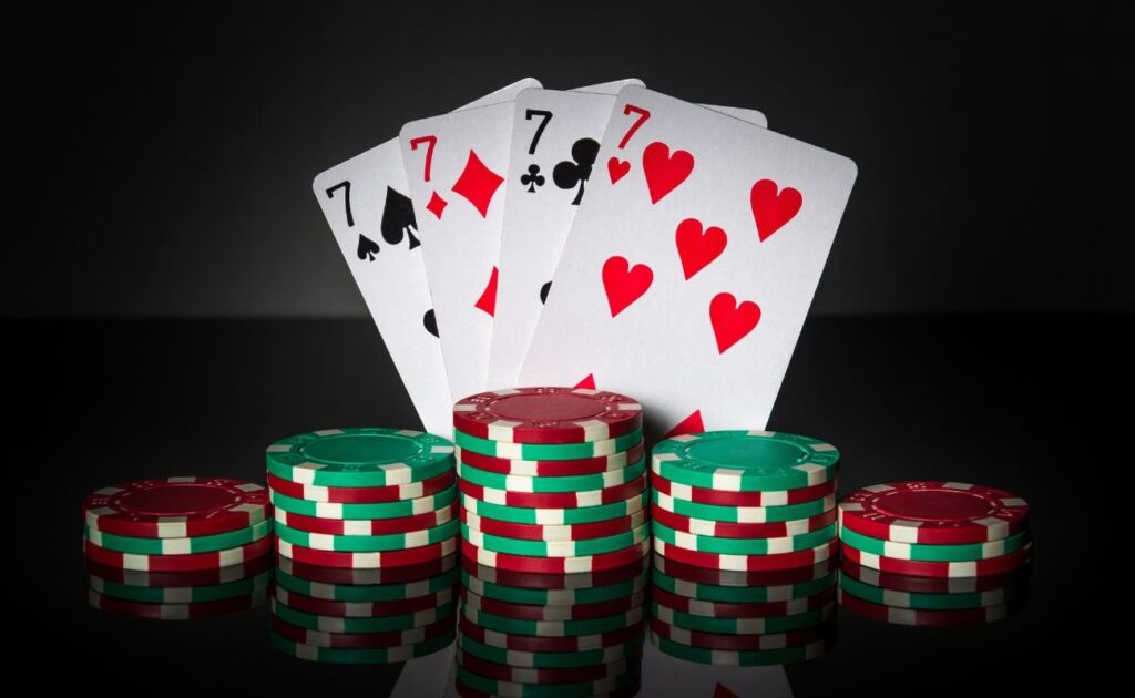 Four of a kind made up of 7s on a reflective black table, with a black background and small stacks of poker chips in the foreground.