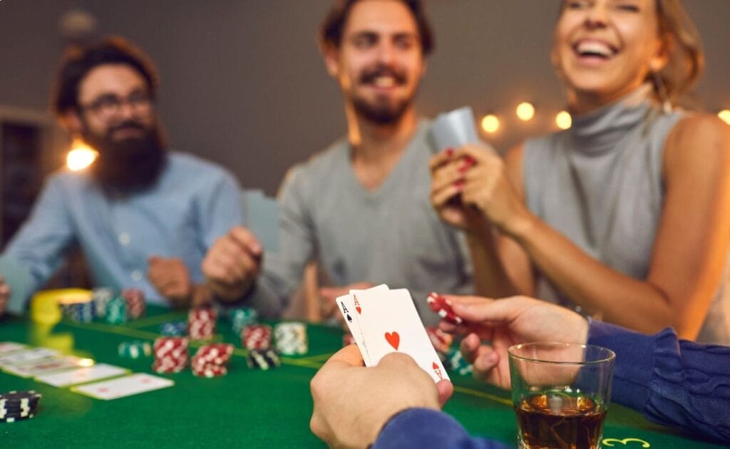 Man holding an Ace of Spades and an Ace of Hearts in one hand, a poker chip in the other, at a poker table. Three other players smiling and laughing in the blurred background.