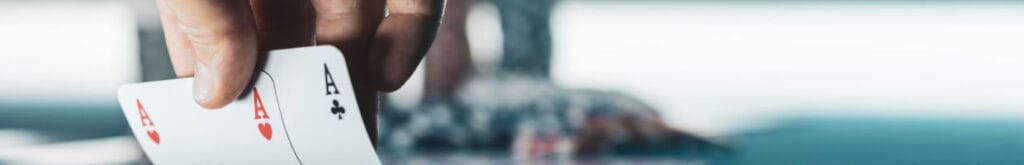 Hand lifting a pair of cards to reveal an ace of hearts and an ace of clubs. Stacks of poker chips in the blurred background.