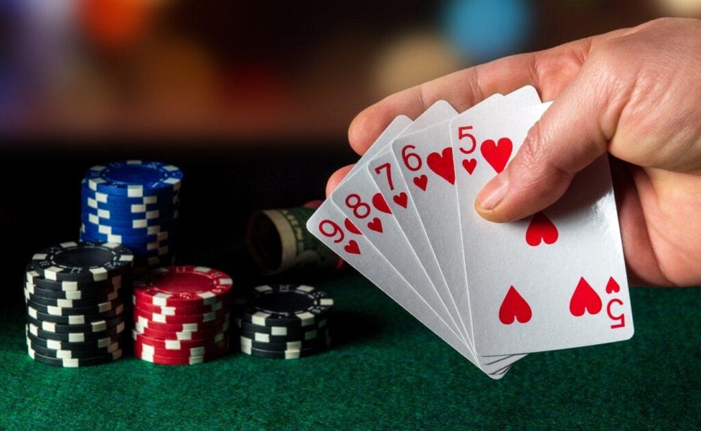 A poker player holds a straight flush. There are stacks of chips and a roll of cash in the background.