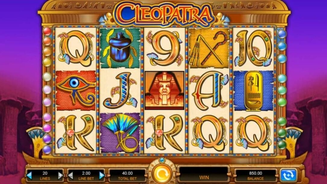 A screenshot of the slot reels in the Cleopatra slot. The background features a gradient purple sky with shadowy pillars and lotus columns, evoking ancient Egyptian architecture. In the foreground are the slot reels with a five-reel layout in a gold Egyptian-style frame with colored orbs on the sides. The game’s logo is visible at the top center of the reels. Each reel contains a variety of themed symbols, including the Eye of Horus, scarabs, scepters, fans, hieroglyphic tablets, and regular slot symbols, like the A, K, Q, and J in an Egyptian style. Below the reels is the game interface.