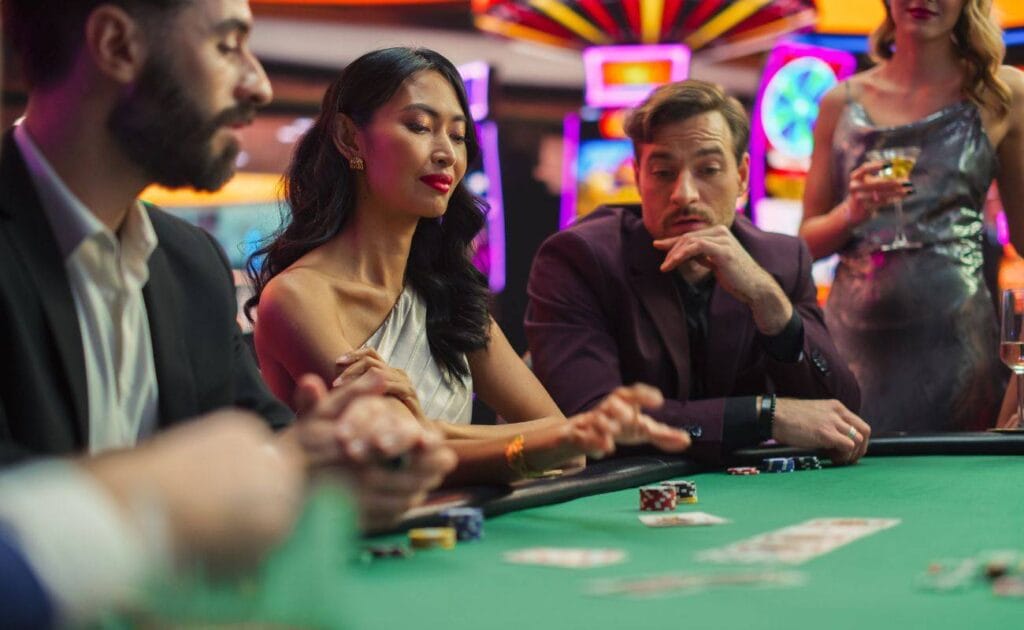 People sitting around a poker table, in a casino, engaging in a game of poker, with poker chips and playing cards arranged on the table. 