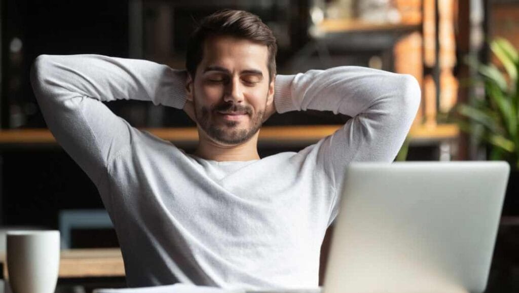 A person sitting at their desk with a laptop open and their hands on the back of their head, looking relaxed.
