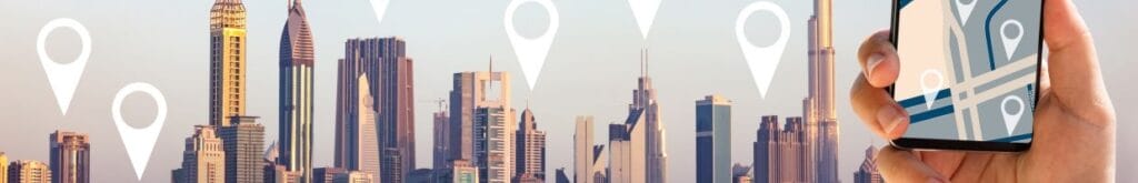 A hand holding a phone showing a GPS in the foreground, and a cityscape in the background with location pins above the buildings.