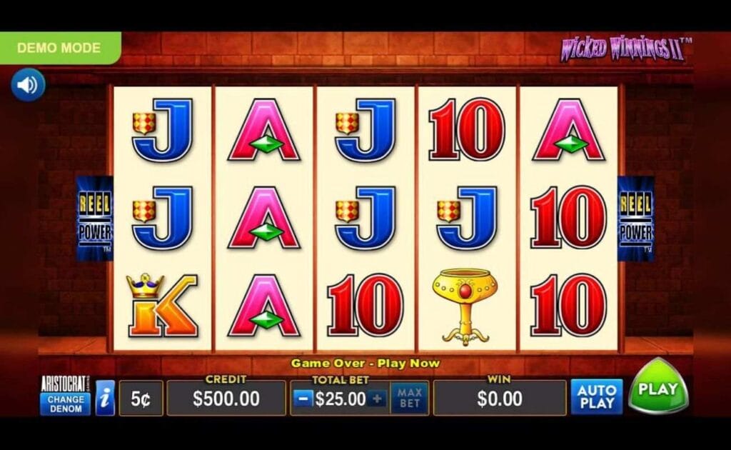 A screenshot of the slot reels in Wicked Winnings II. The game is set against a brick wall with a reddish tinge. The reels contain a variety of symbols, including a chalice, A, K, Q, and 10. The Reel Power logo is on either side of the reels.