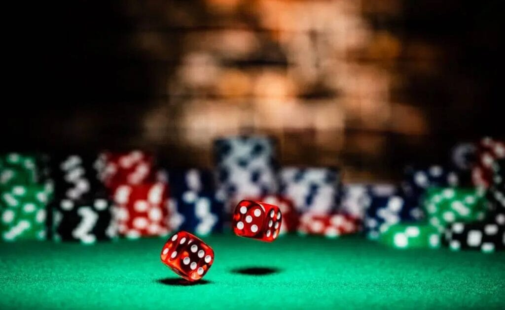 A pair of red dice rolling on a green casino table with blurry stacks of casino chips in the background.