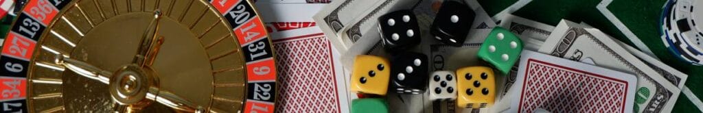 A range of casino game equipment sitting on a casino game table, including a mini roulette wheel, dice, playing cards, and casino chips.