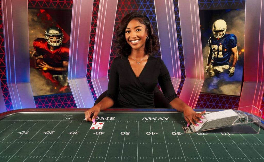 The U.S. version of the Football Studio live dealer game. A game show host reveals cards on a table that looks like an American football field. There are two football players in red and blue uniforms on the studio background. 