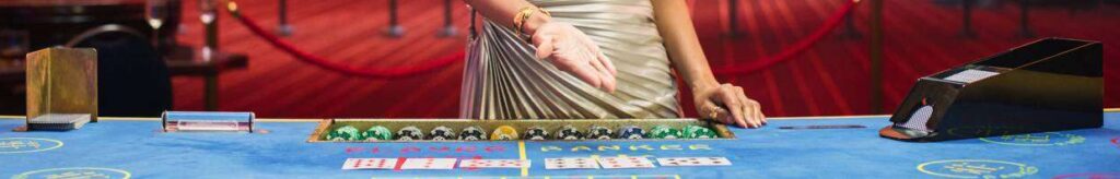 A female casino dealer in a silver dress gestures toward a baccarat table.