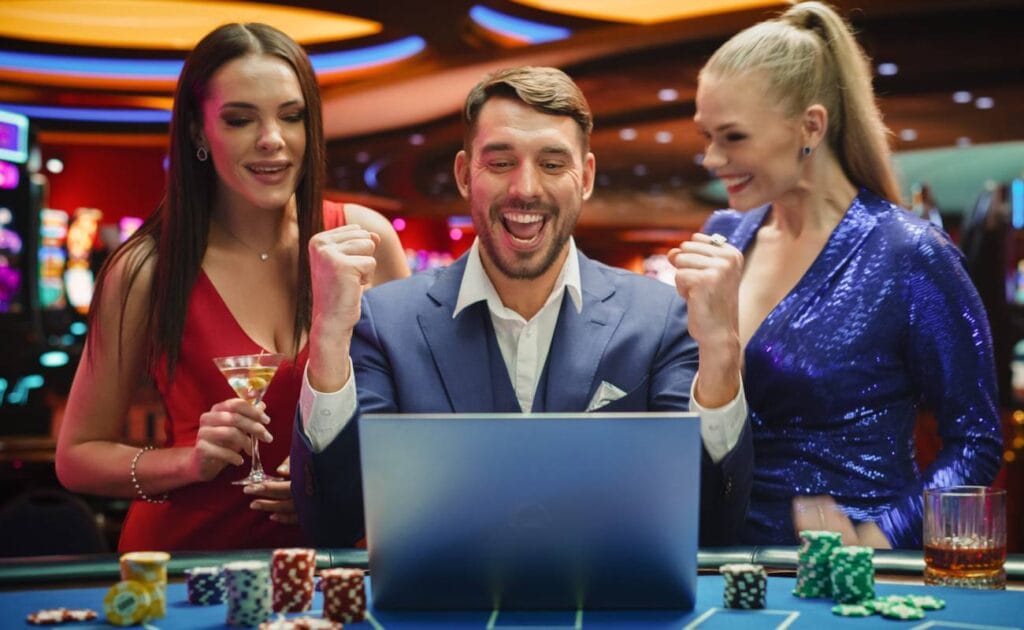 A man celebrates in front of a laptop on a casino game table. There are stacks of casino chips and a drink on the casino game table. One woman in a red dress and another in a blue dress stand on either side of him, smiling.