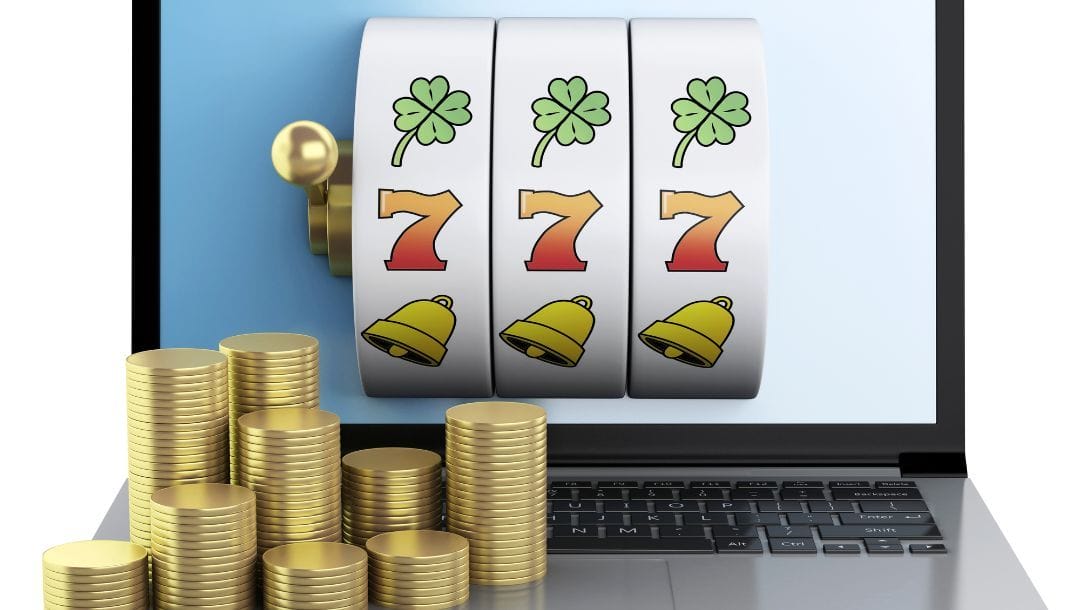 Slot machine reels displayed on a laptop screen showing clovers, red sevens, and golden bells, with stacks of gold coins in the foreground.