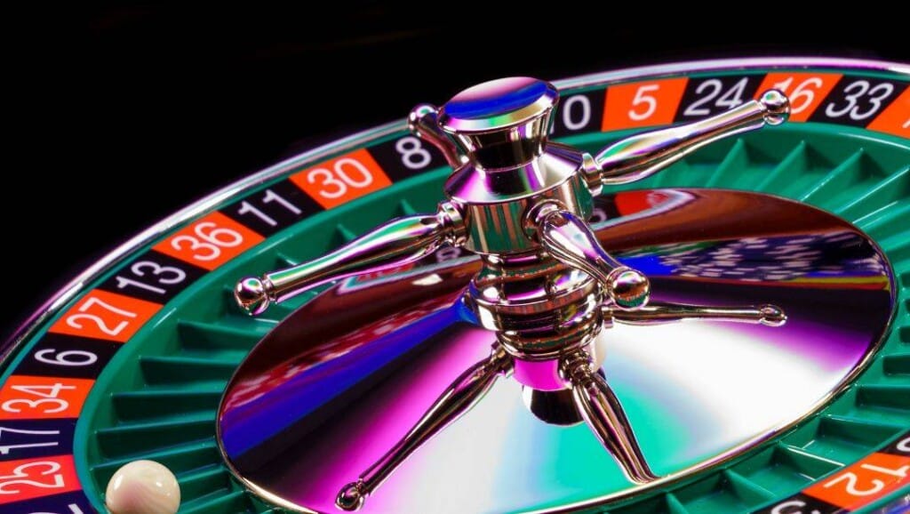 A close-up of a colourful roulette wheel mid-spin, with the white ball landing near the red and black numbered pockets under vibrant casino lights.