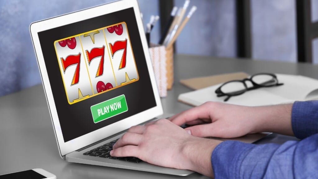 Person playing an online slot game on a laptop showing three 7s and a green “Play Now” button, with stationery and glasses on the desk.