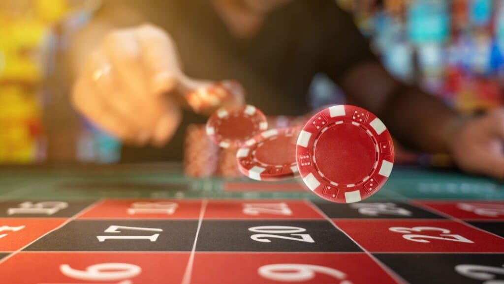 Red casino chip flying through the air toward a roulette table during a game, with a player in motion and a blurred casino background.