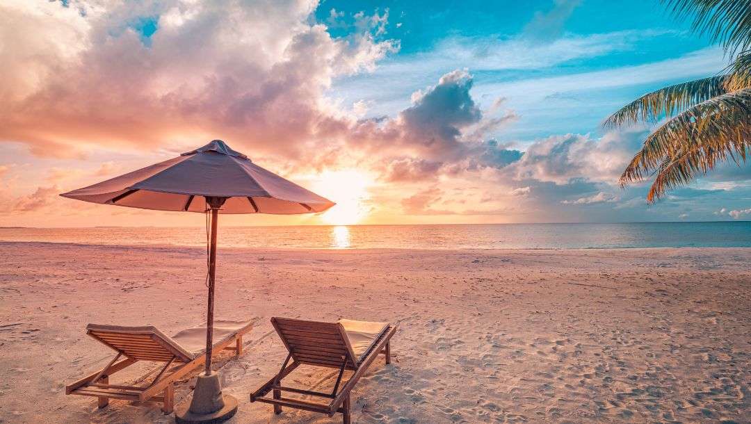 Two lounge chairs and an umbrella on a tropical beach at sunset.