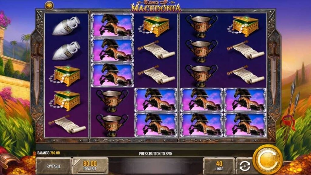 King of Macedonia base game screen showing the main reel grid framed in ornamental metalwork with the game controls set on stone blocks. The background features an oil painting-like landscape at sunrise, picturing a Macedonian villa in a lush valley between rolling hills.