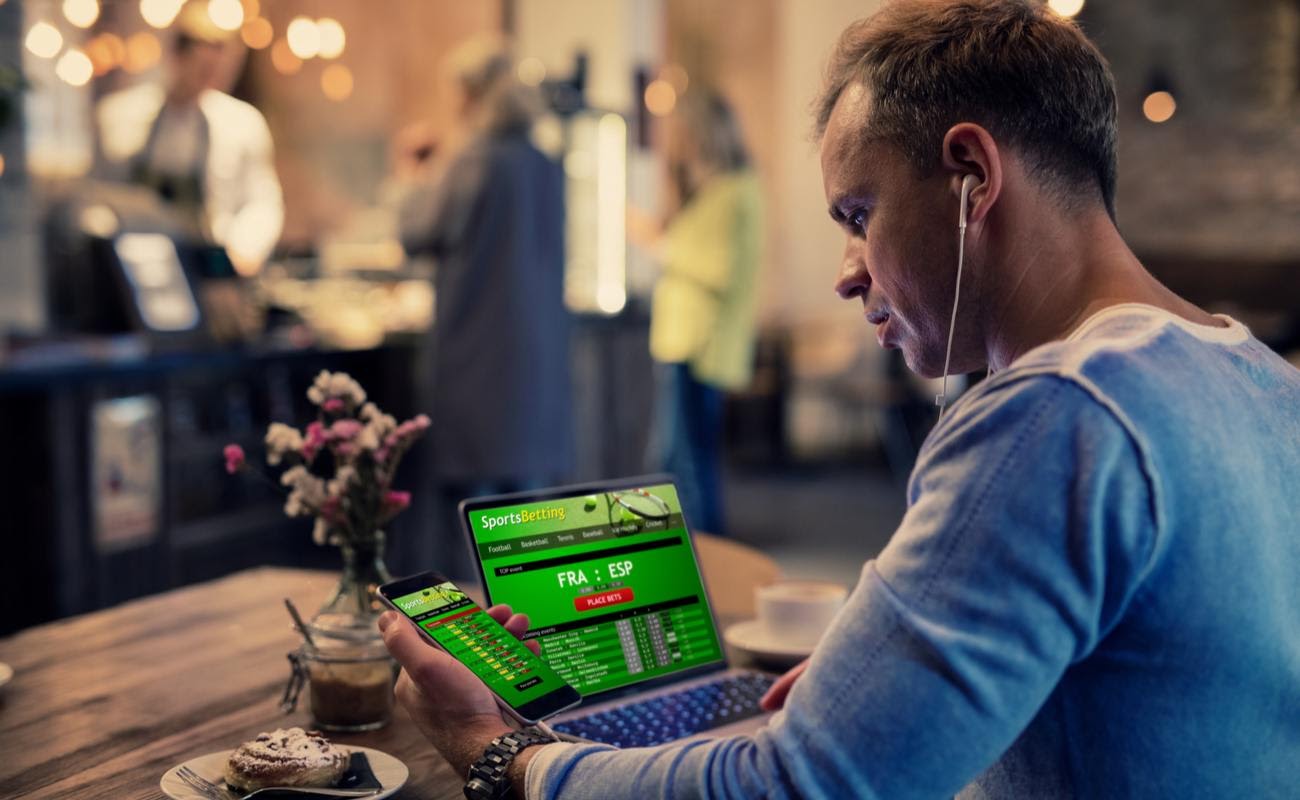A man in a cafe looking at sports betting results on his laptop and smartphone