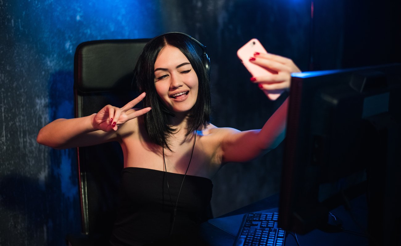 A girl sitting at her desk taking a selfie with her phone posing with a peace sign and winking.