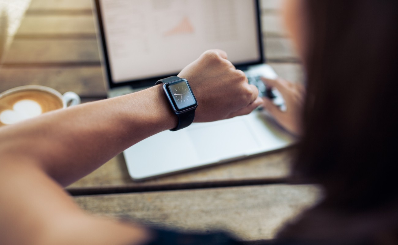 Close up shot of woman checking the time on her smartwatch with laptop and cup of coffee in the background