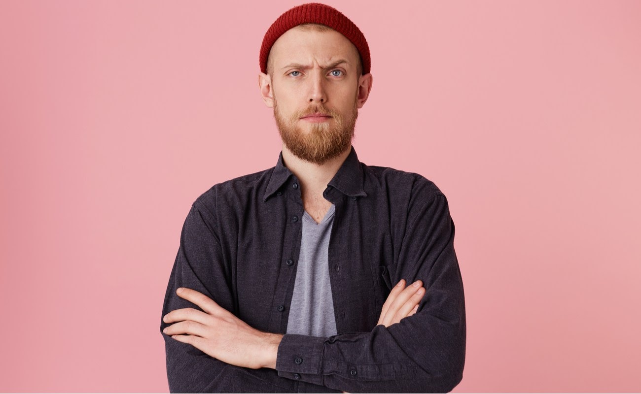 Close up of bearded man standing in front of pink background, looking at the camera with one raised eyebrow