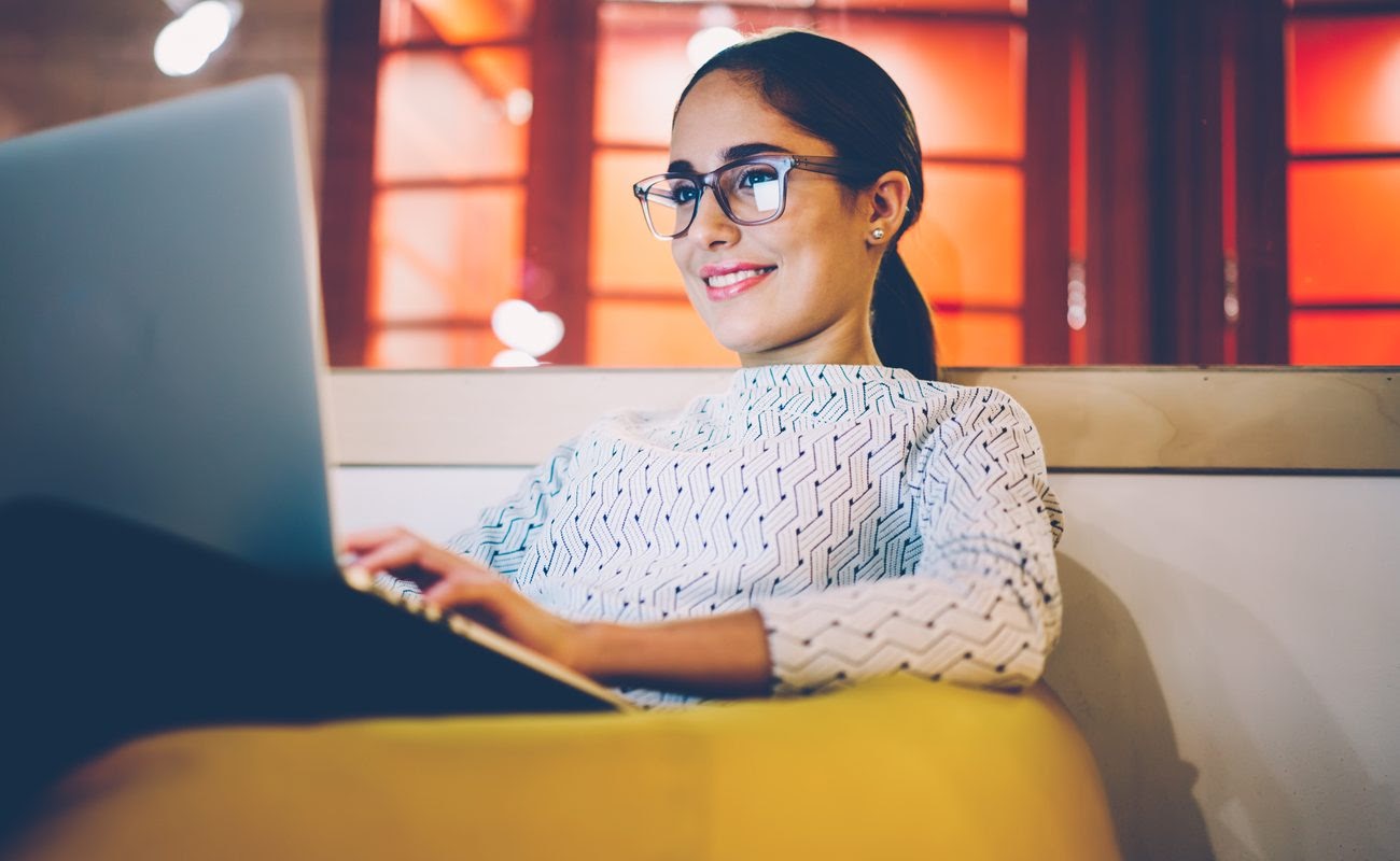 Smiling woman in eyewear looking at a laptop on the sofa