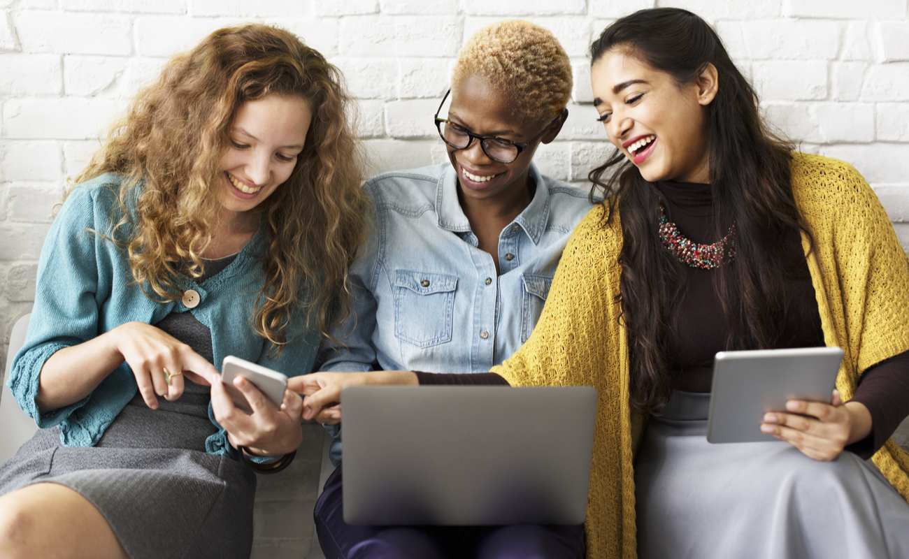 Three female friends laughing as they share screens from their technological devices including laptop android and tablet