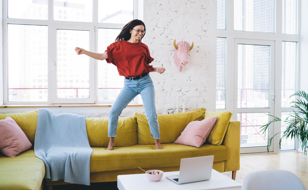 Young joyful woman dancing on yellow couch in living room near coffee table with laptop