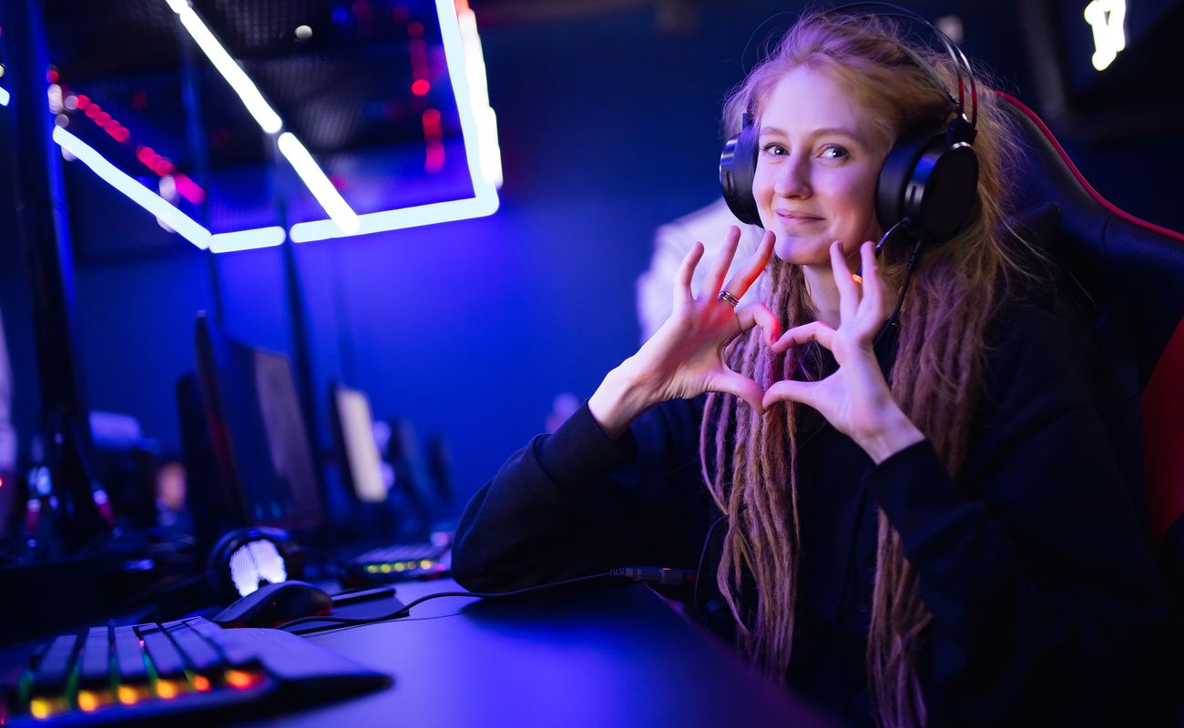 A gamer wearing headphones makes a heart symbol with her fingers while sitting in front of her PC.