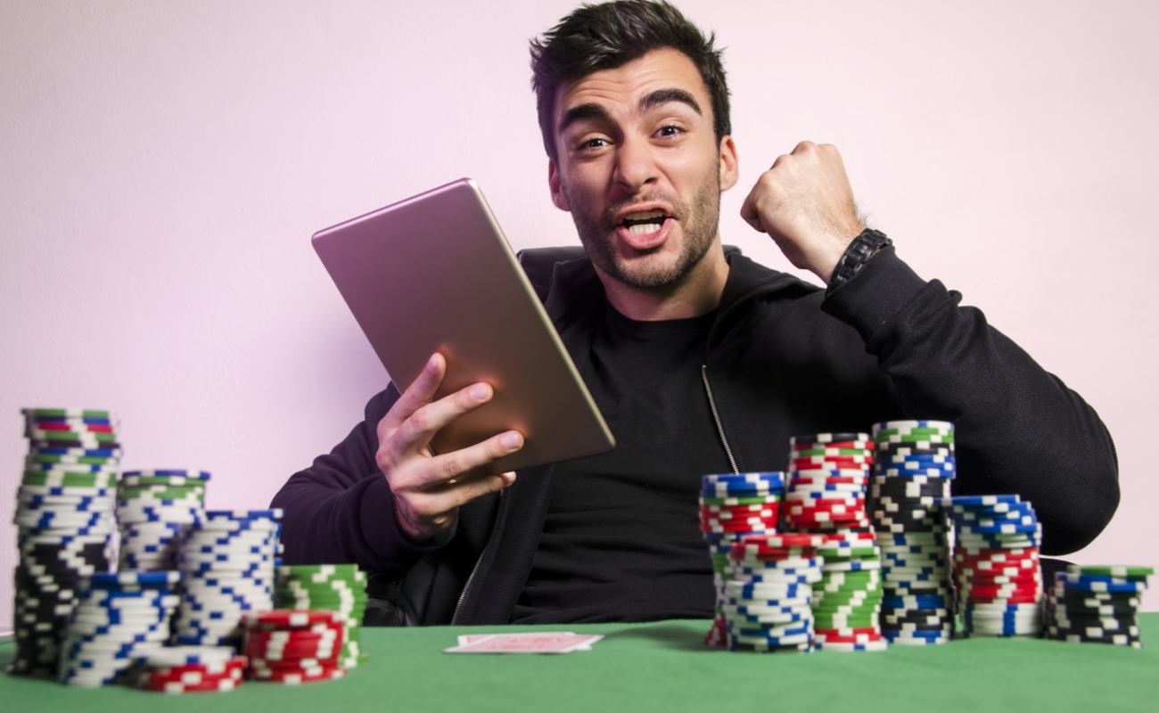 A young man celebrates his online slot win on his tablet, while sitting at a table with casino chips and playing cards.