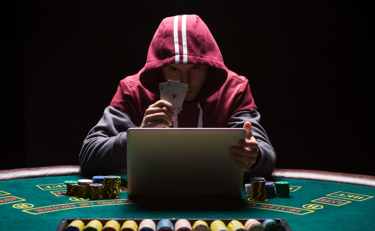 Online poker player sitting at a laptop on a casino table wearing a red hoodie and holding a pair of aces.